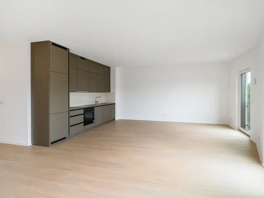 Open-plan kitchen with tall gray cabinets along left wall, built-in oven and sink, light wood floor, white walls, and a glass door to the balcony.
