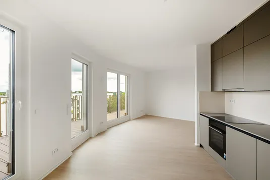 Bright open-plan living area with light wood floors, curved white walls, and large glass doors to a balcony; sleek gray kitchen with built-in oven.