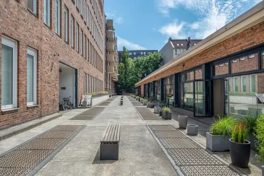 Paved courtyard between red-brick buildings with benches, planters, metal grates and parked bicycles under a bright blue sky.