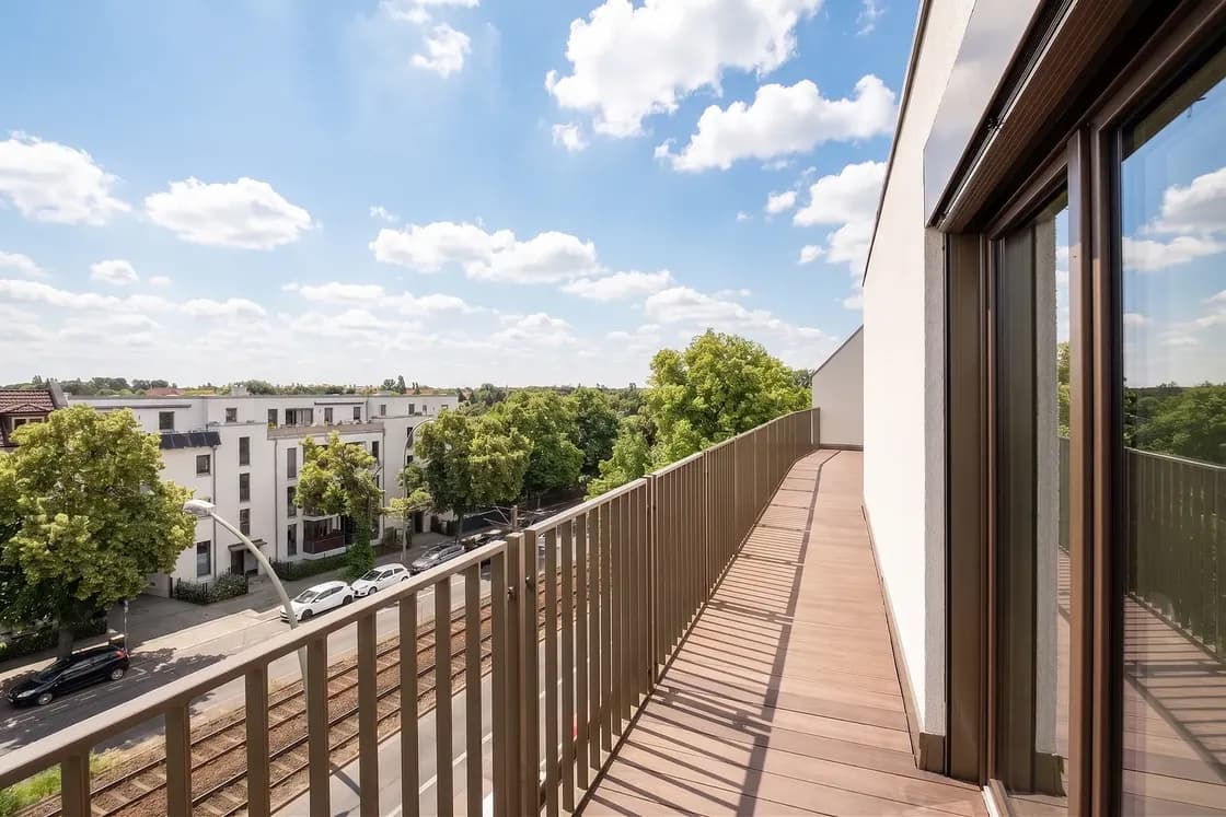 Balcony view from a modern building: wooden deck, brown railing, glass doors, overlooking white apartment blocks, trees, a railway, under a bright blue sky. AI-generated image
