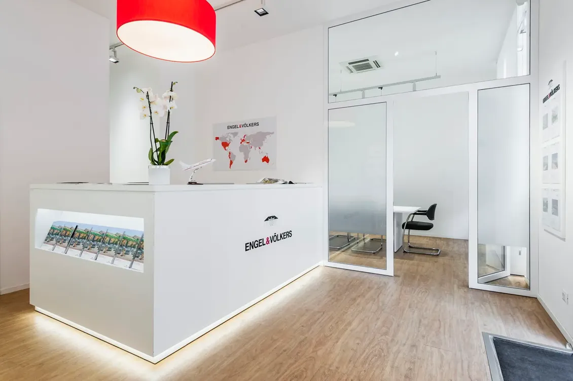 Luminosa reception immobiliare in stile minimalista con scrivania bianca Engel & Völkers, un'orchidea, un modellino di aereo, una lampada a sospensione rossa e una sala riunioni in vetro.