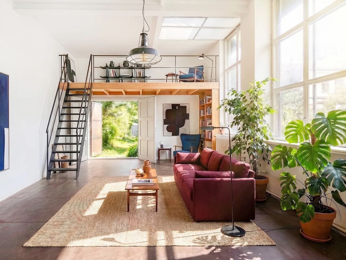 Sunlit modern loft living room with a burgundy sofa, large windows, potted plants, woven rug and a wooden mezzanine reached by metal stairs.