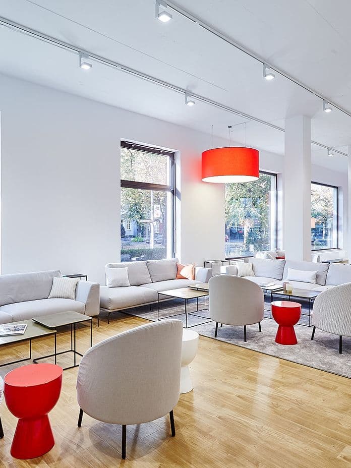 Modern lounge with white sofas, round chairs, red accent tables, and large red ceiling lamps. Bright windows and wooden flooring.