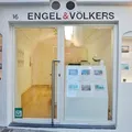 Vetrina bianca dell'agenzia immobiliare Engel & Völkers con porta in vetro, display illuminati per gli annunci immobiliari nelle nicchie laterali e su un espositore frontale.