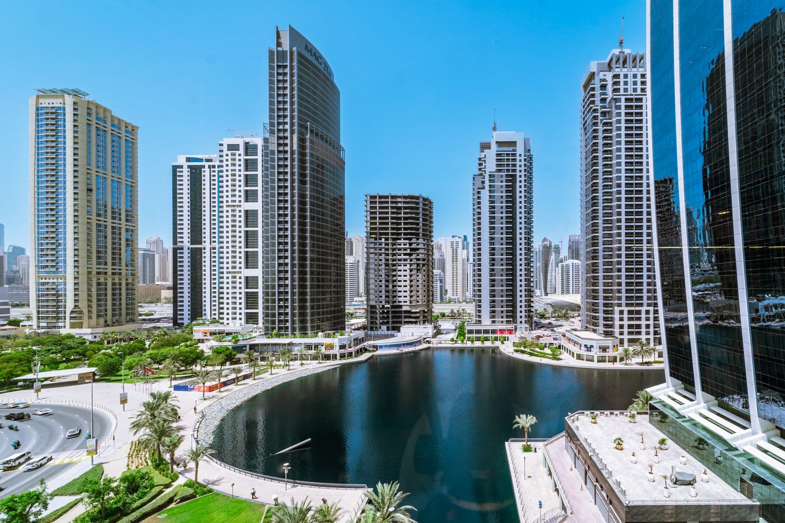 Skyline view of modern high-rise buildings surrounding a circular water feature, with a clear blue sky above and greenery below.