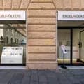 Vetrina di Engel & Völkers in una facciata di pietra color biondo/tan, con bacheca espositiva che mostra gli annunci immobiliari e una porta di vetro aperta con un porta depliant