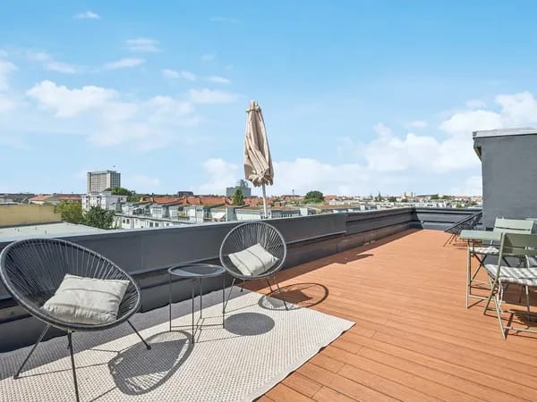 Rooftop terrace with wooden deck, two round black wicker chairs with cushions, a closed beige umbrella, a small table, and distant city skyline under a blue sky.
