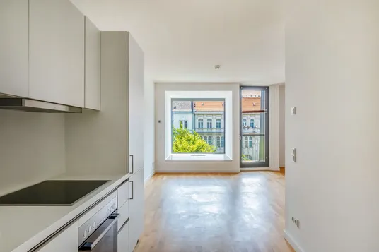 Bright empty modern studio apartment with white kitchenette on left, light wood floor, large square window framing tree and ornate building.