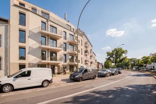 Modern beige apartment building with balconies along a curved street; several vans parked on the curb; blue sky overhead. AI-generated image