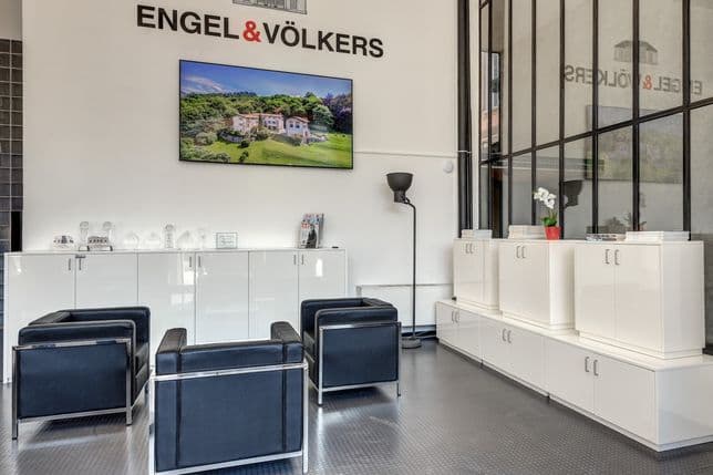 Bright Engel & Völkers real estate office in Lake Como with a logo wall, a screen displaying a countryside estate, white cabinets, and blue chairs.