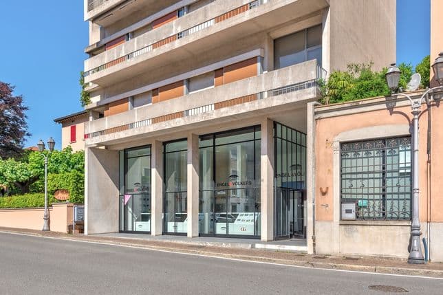 Street view of a modern building with a glass storefront on the ground floor and orange balcony accents, beside a peach-colored building and lamps.