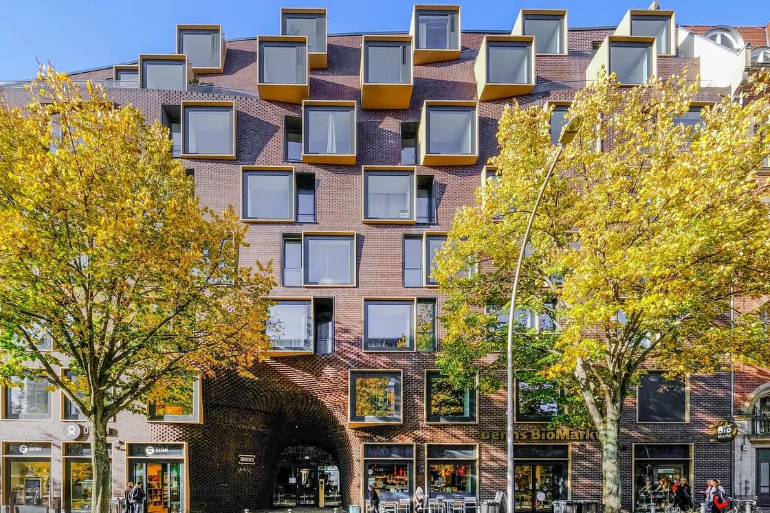 Brick building with protruding box windows, two yellow-leaved trees in front, ground-floor shops and pedestrians.