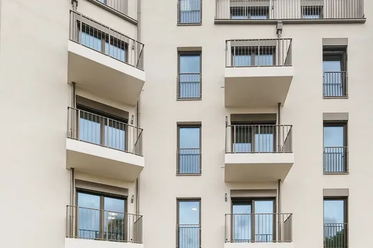 Modern beige apartment building with multiple stacked balconies, metal railings, and large windows; central ground-floor entry opening to a courtyard.