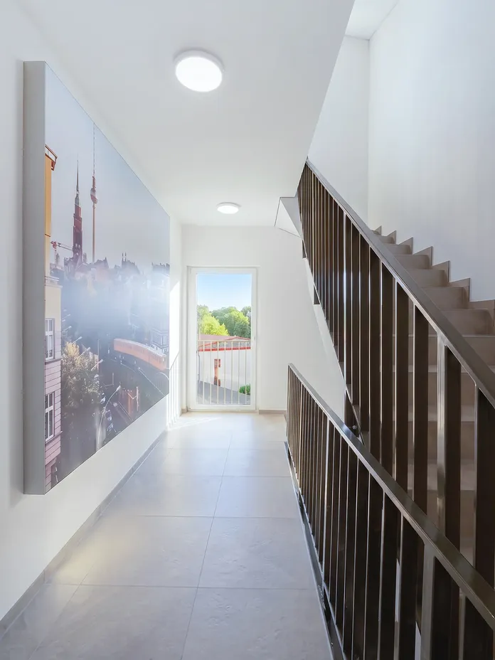 Bright modern hallway with a dark metal staircase on the right, a large cityscape mural on the left, and a window at the end overlooking trees.