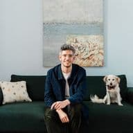 Ένας άνδρας με γυαλιά κάθεται σε έναν πράσινο καναπέ δίπλα σε ένα άσπρο σκύλο σε ένα γαλάζιο δωμάτιο με έναν πίνακα.