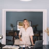 Μια χαμογελαστή γυναίκα με μπεζ φόρμα στέκεται σε ένα γραφείο με ένα λάπτοπ και λουλούδια σε ένα μπλε δωμάτιο.