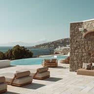 Poolside terrace with beige loungers beside a stone wall, an infinity pool, and a sea view over a hillside village.
