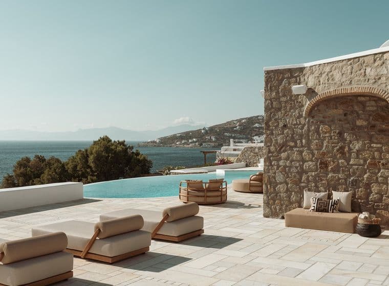 Poolside terrace with beige loungers beside a stone wall, an infinity pool, and a sea view over a hillside village.