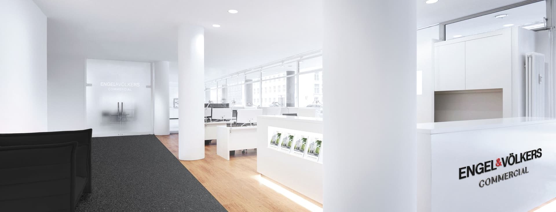 Modern real estate office interior with sleek white desks, computers, glossy reception desk, and a black couch on a carpeted floor.