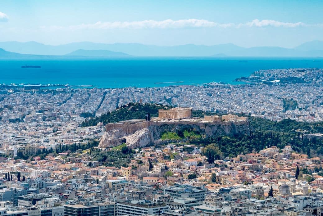 Athens cityscape with the Acropolis on a hill, viewed from above. The Aegean Sea and distant mountains are in the background.