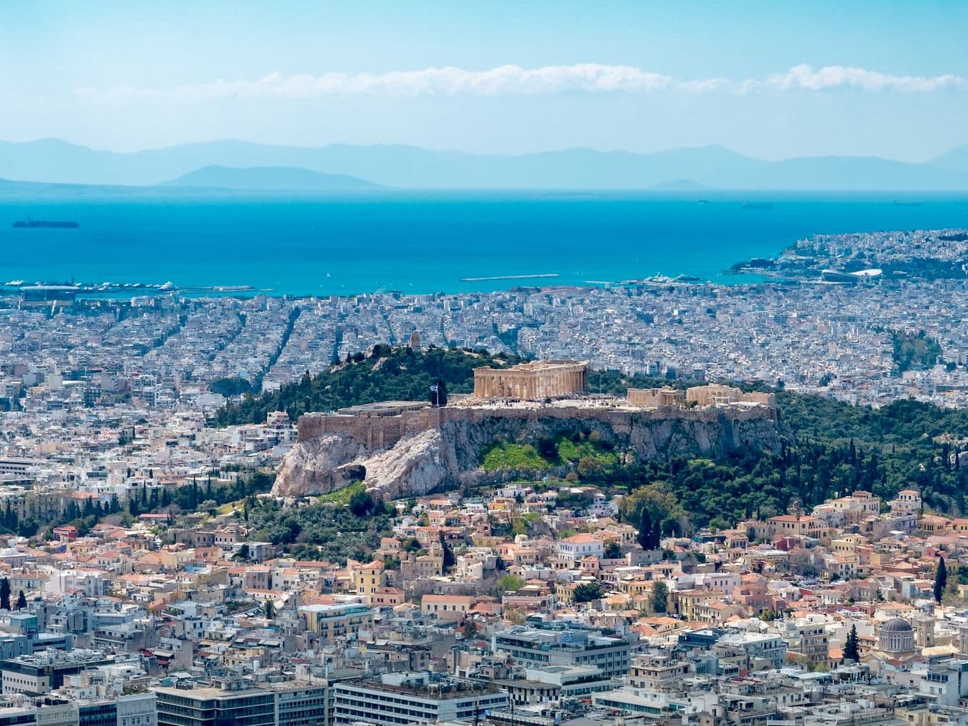 Athens cityscape with the Acropolis on a hill, viewed from above. Blue sea and mountains in the background.