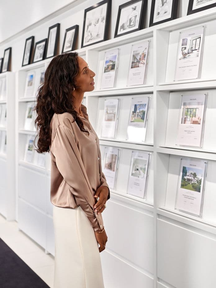 A woman in a satin blouse and white skirt looks at framed property listings on a white wall in a modern real estate office.