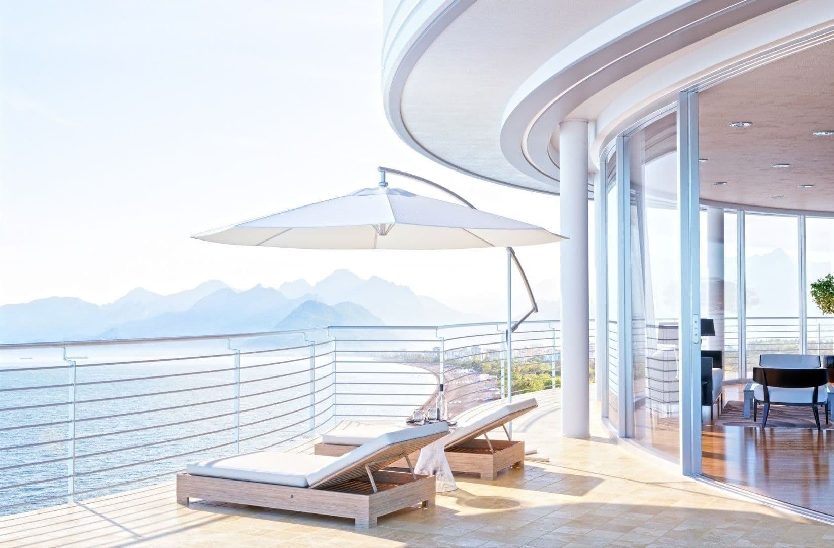 Balcó de luxe amb vista sobre l'oceà. Dues gandules sota un paraigua blanc. Portes corredisses de vidre condueixen a un interior elegant.
