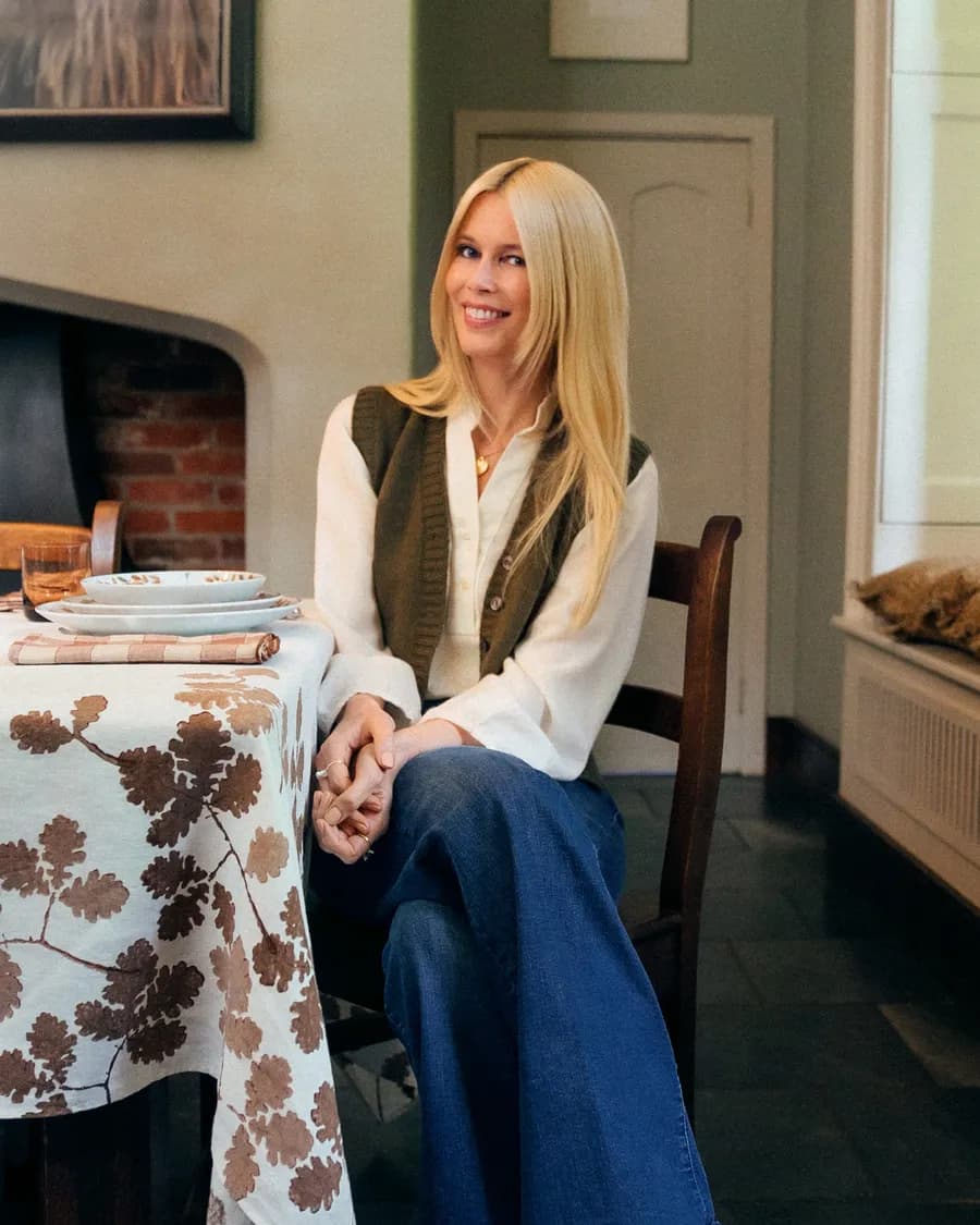 A smiling blonde woman in a white blouse and an olive-green waistcoat is sitting at a dining table with a floral tablecloth and crockery; a dog is resting beside her.