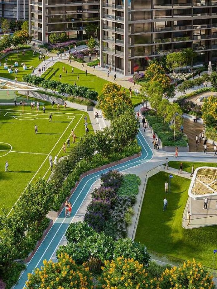 Aerial view of a modern residential complex with landscaped gardens, a soccer field, walking paths, and recreational areas surrounded by tall buildings.