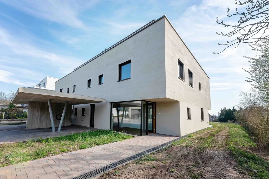 Modern white two-story building with flat roof, glass entry under a concrete canopy supported by angled columns, and small square windows.
