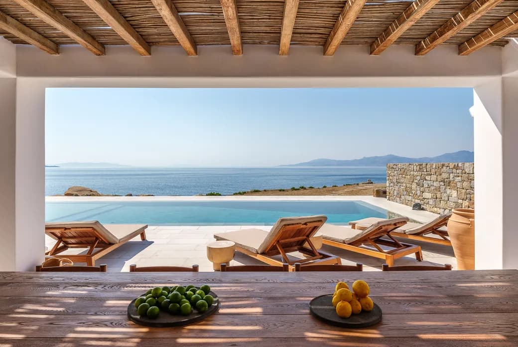 Open-air terrace with a wooden beam roof shows a pool and sea beyond; lounge chairs by the pool and a stone wall, fruit bowls on a wooden table.