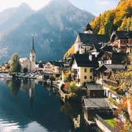 Scenic view of a lakeside village with colorful houses, a church, and autumn trees, set against a backdrop of mountains and a calm lake.
