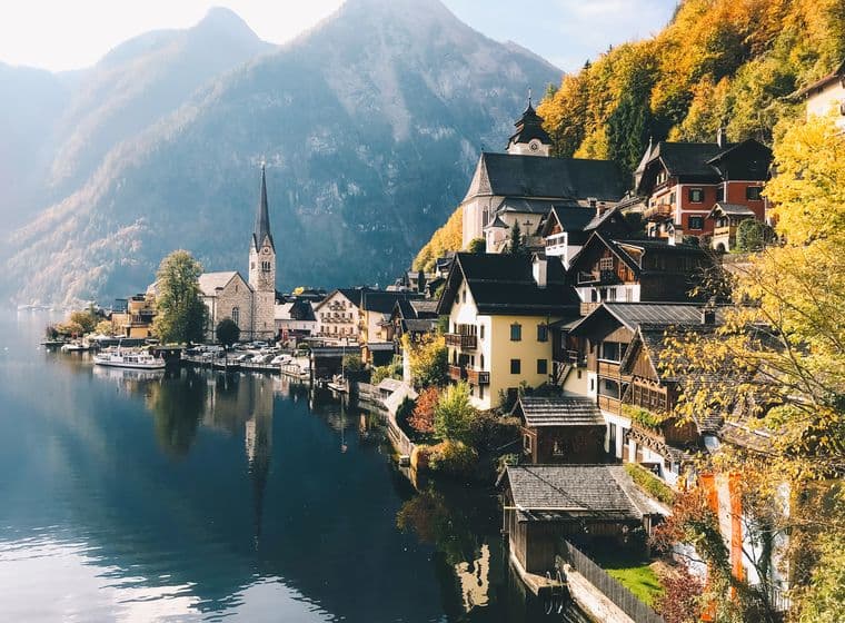 Scenic view of a lakeside village with colorful houses, a church, and autumn trees, set against a backdrop of mountains and a calm lake.