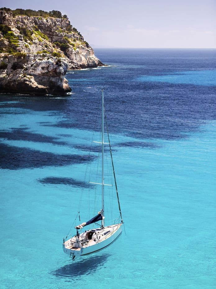 A sailboat floats on clear turquoise water near a rocky cliff under a bright blue sky.