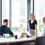 A group of professionals are in a meeting room with large windows. A woman stands, smiling, while others sit at a table with laptops.