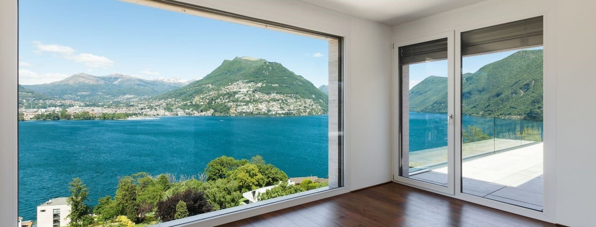 Habitació lluminosa amb terres de fusta i una gran finestra amb vista a un llac blau i muntanyes.