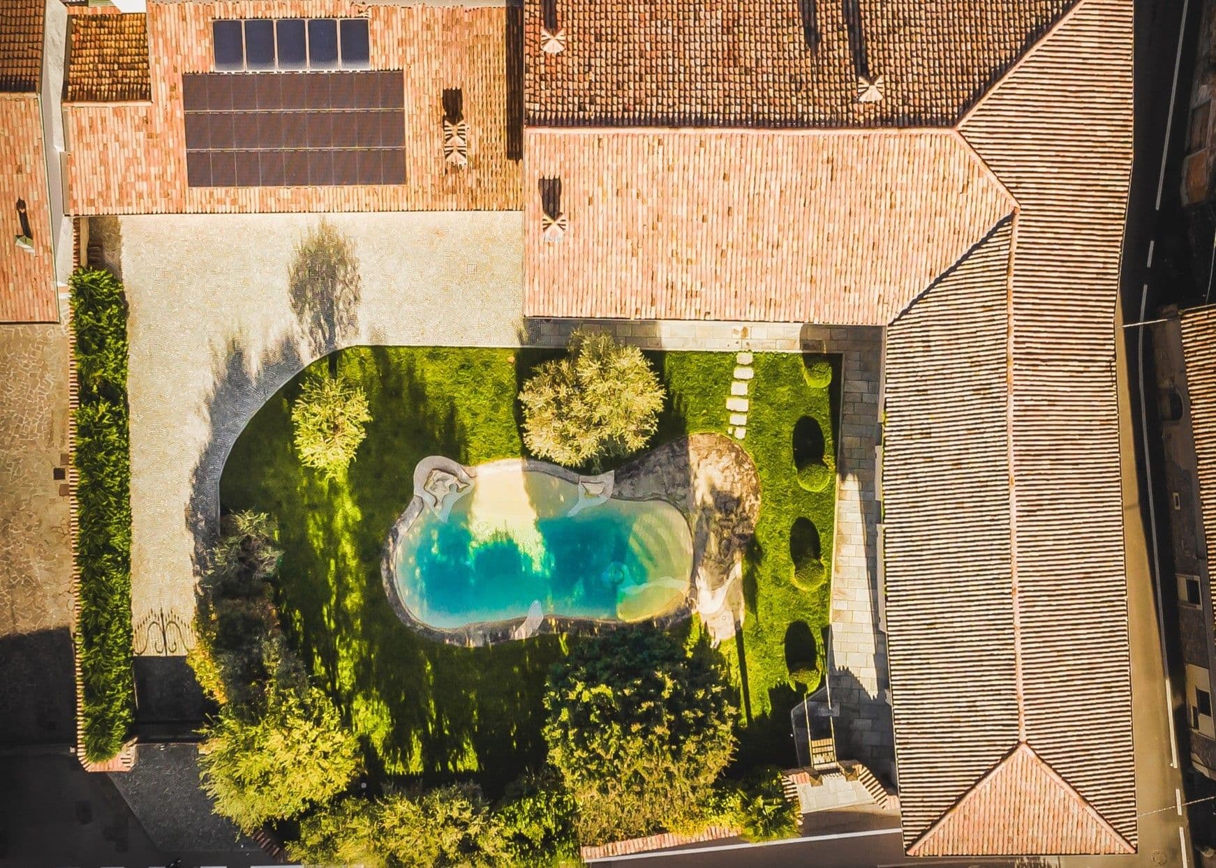 Aerial view of a house with a red-tiled roof, solar panels, a lush green garden, and an irregularly shaped swimming pool.