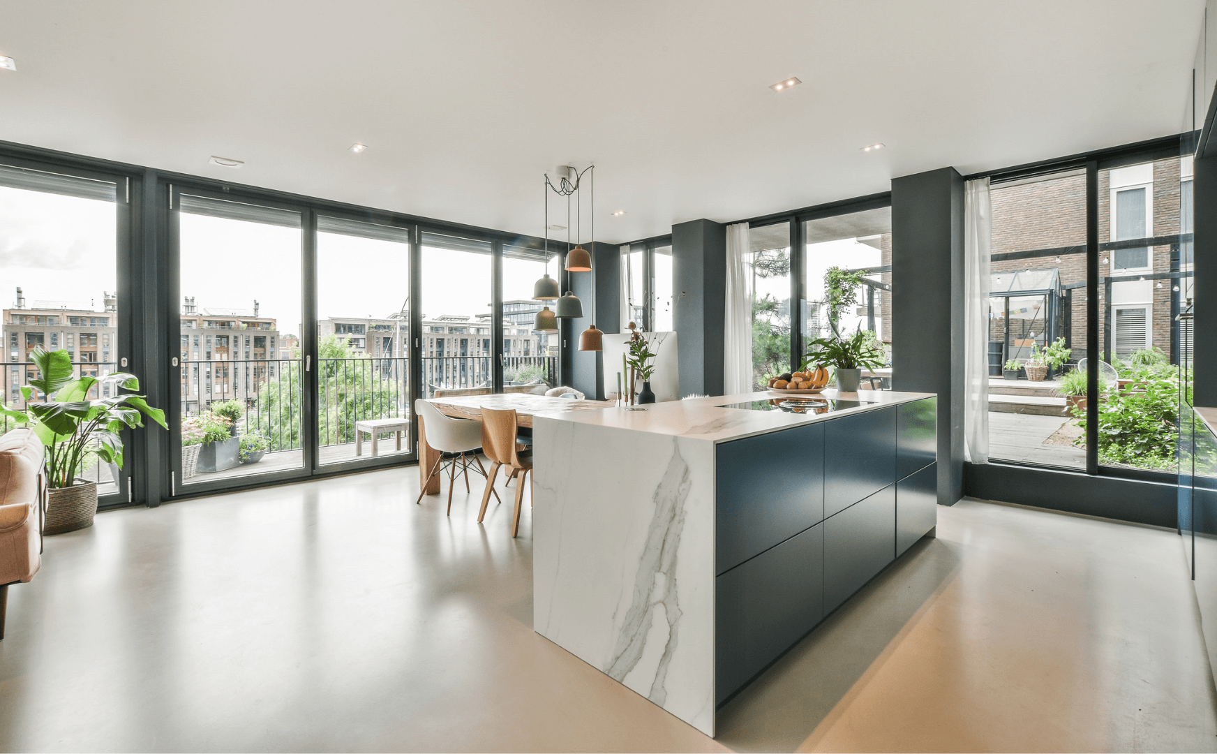 Cuina lluminosa i moderna amb grans finestrals. S'hi veu una illa de marbre i una taula de fusta amb cadires. A l'exterior, hi ha una vista d'una ciutat i un jardí.