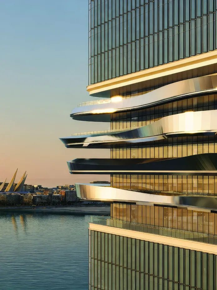 Modern glass building with stacked curved balconies overlooking a calm river at sunset.