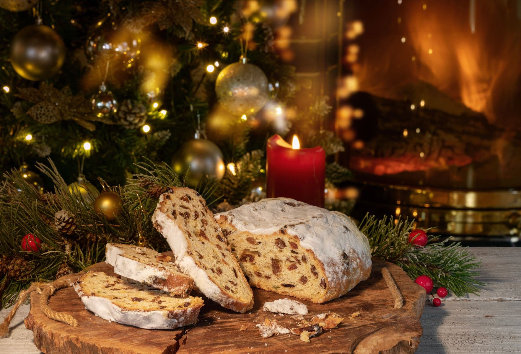 Aufgeschnittenes Stollenbrot auf einem Holzbrett, mit einer brennenden roten Kerze und Weihnachtsdekoration im Hintergrund.