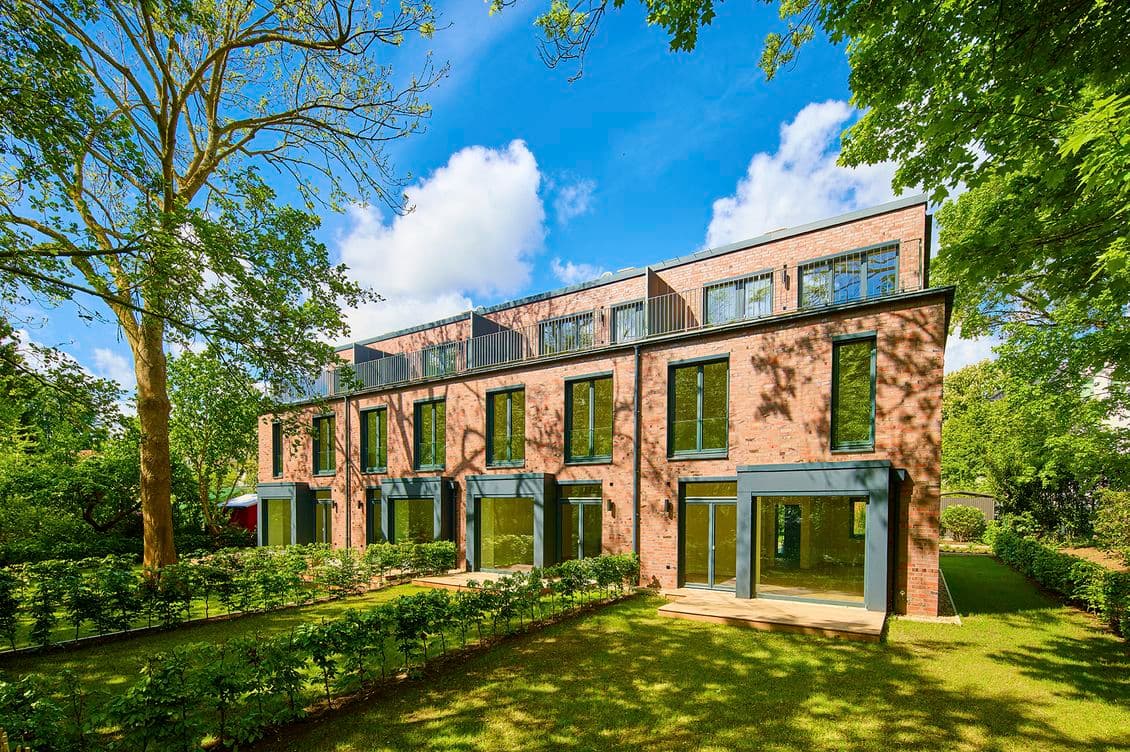 Ein modernes, dreistöckiges Reihenhaus aus Backstein mit großen Fenstern, eingebettet in einen üppigen, grünen Garten unter einem klaren blauen Himmel.