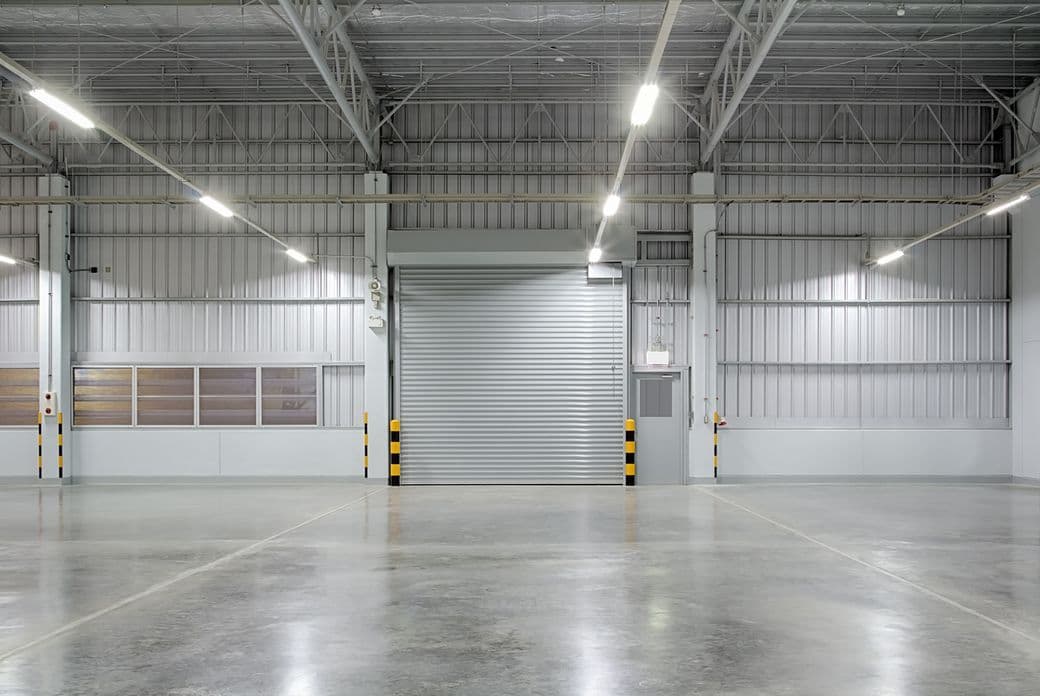 Empty industrial warehouse with polished concrete floor, overhead lighting, metal walls, and a large closed roller shutter door.