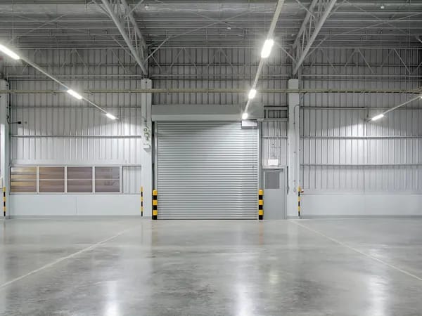 Empty industrial warehouse with polished concrete floor, overhead lighting, metal walls, and a large closed roller shutter door.