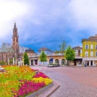 Una vivace piazza cittadina con fiori colorati, edifici storici, una statua e una chiesa, sullo sfondo di montagne e di un cielo nuvoloso.