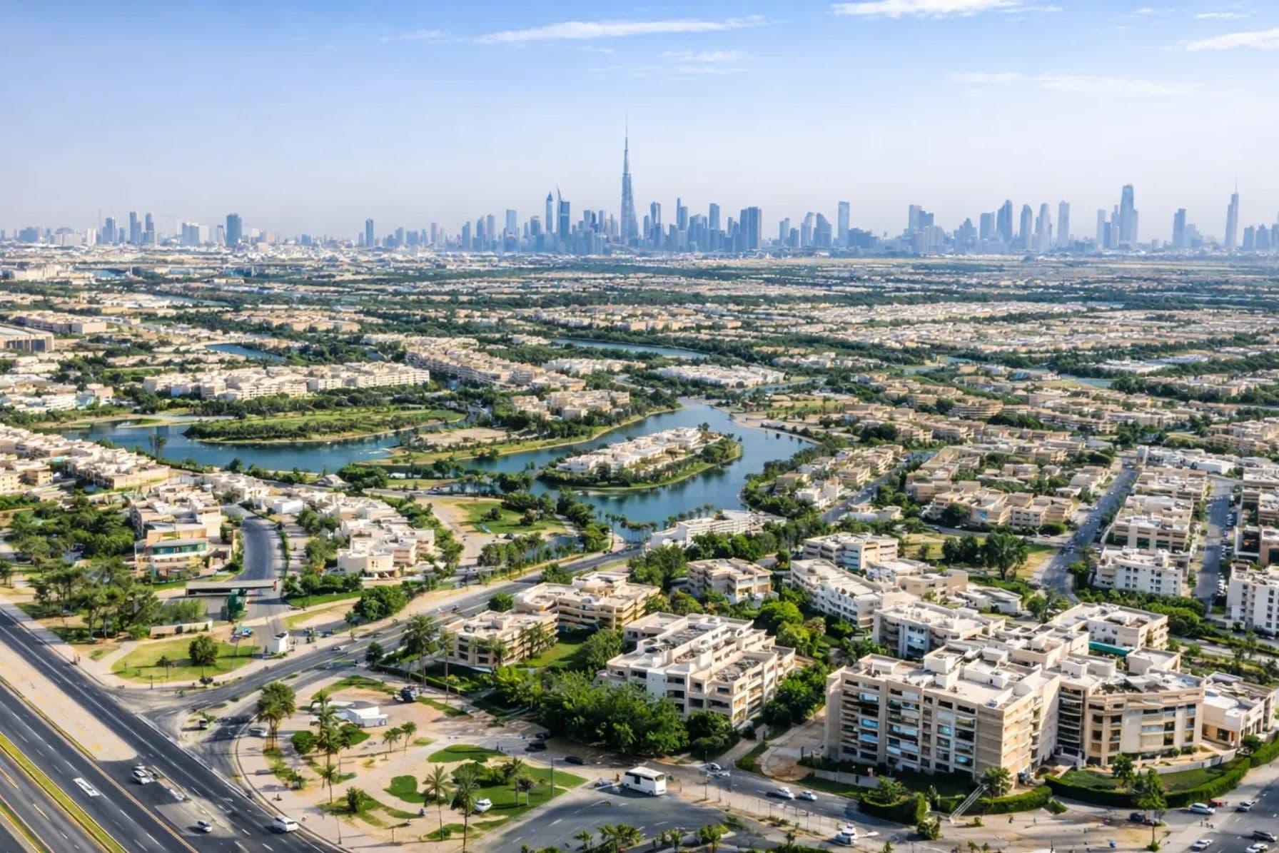 Aerial view of a green suburban area with lakes and roads, leading to a distant city skyline featuring towering skyscrapers under a blue sky.