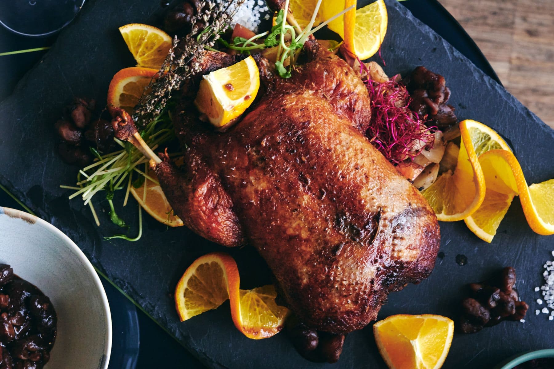 Gebratene Ente, garniert mit Orangenscheiben und Kräutern auf einer Schieferplatte, umgeben von kleinen Schälchen mit Sauce und Gewürzen.