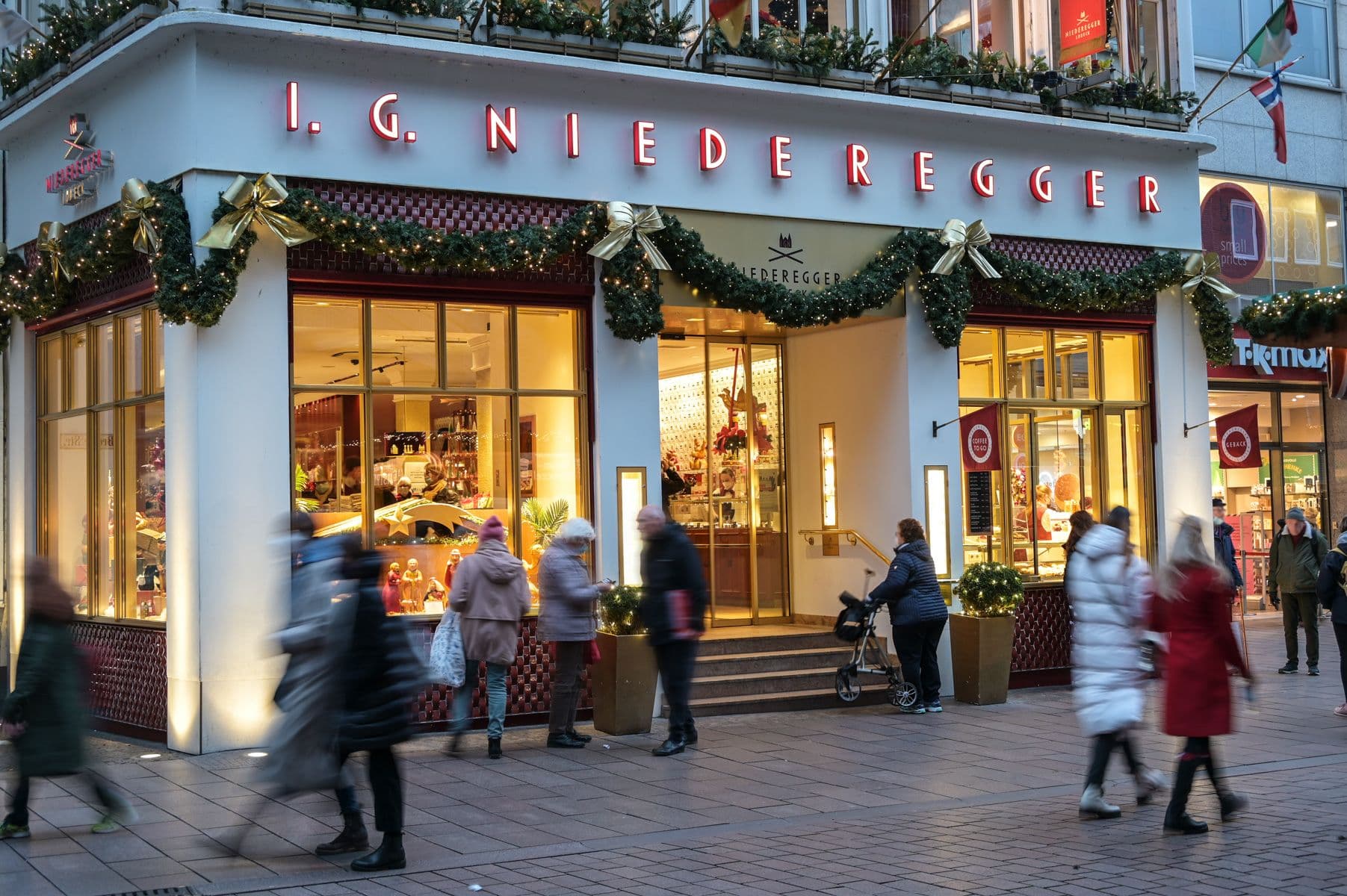Menschen, die auf einer belebten Straße an einem hell erleuchteten, mit Weihnachtsgirlanden und Schleifen geschmückten Geschäft von I.G. Niederegger vorbeigehen.