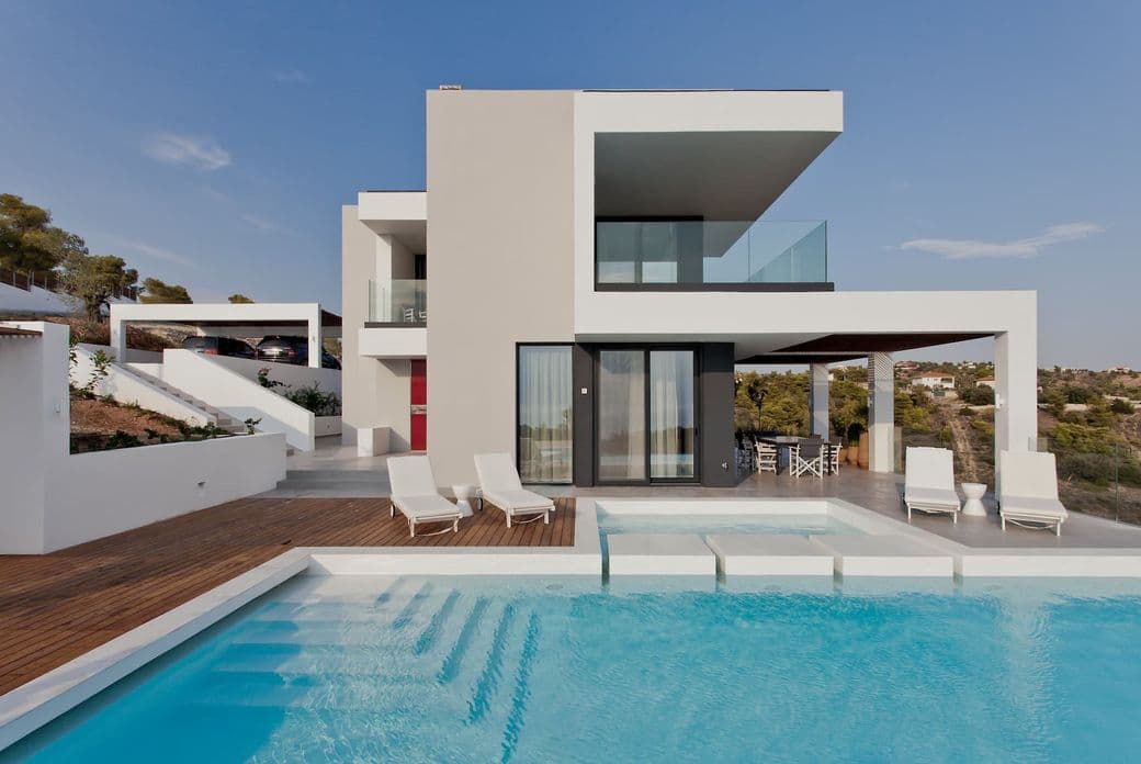 Μοντέρνο διώροφο σπίτι με πισίνα. Λευκές ξαπλώστρες βρίσκονται σε μια ξύλινη βεράντα. Μια κόκκινη πόρτα είναι ορατή στην πλευρά του σπιτιού.