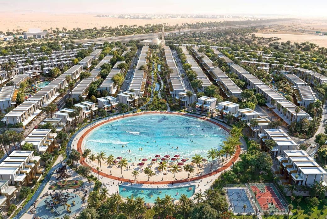 Aerial view of a luxury residential development with a large circular pool, sandy beach, palm trees, playground, and sports courts surrounded by villas.