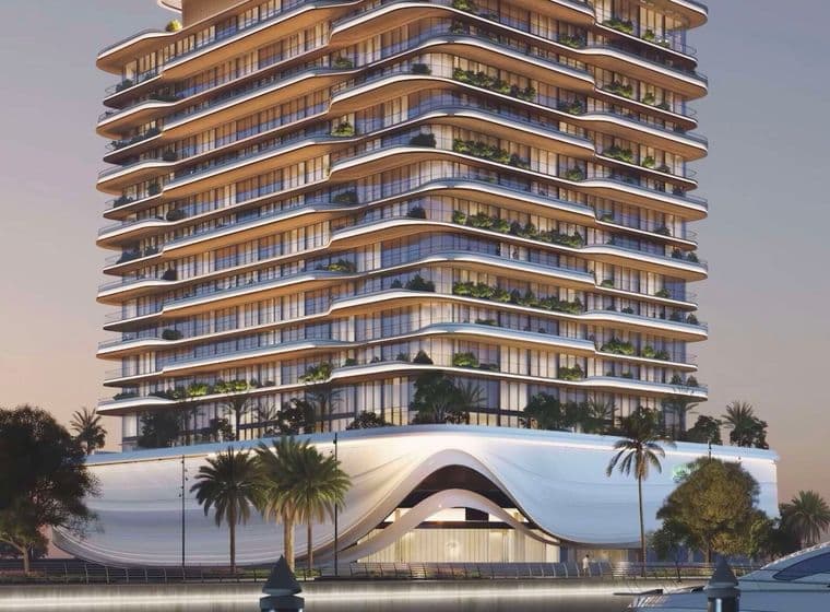 Modern waterfront building with curved balconies, illuminated at dusk. Palm trees line the entrance, and a yacht is docked nearby.